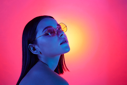 Attractive girl in stylish eyeglasses with black hair and perfect makeup expressing pleasure while relaxing in neon lights