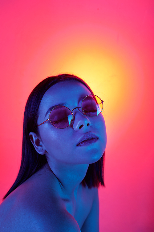 Portrait of sexy girl in stylish glasses keeping eyes closed under blue light, pink and yellow background