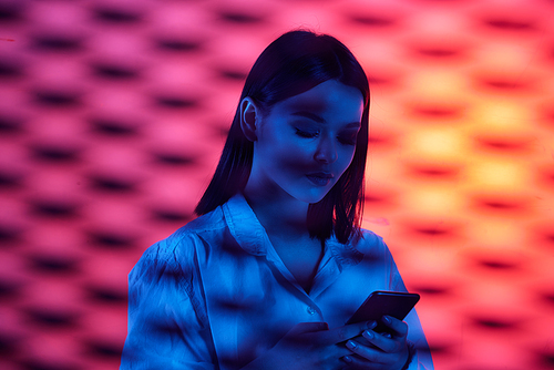 Young attractive female office worker in white shirt scrolling in smartphone on neon background behind bars