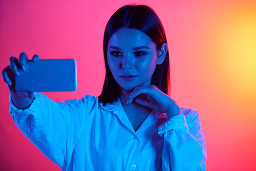 Beautiful young woman in blouse taking selfie on smartphone in neon light