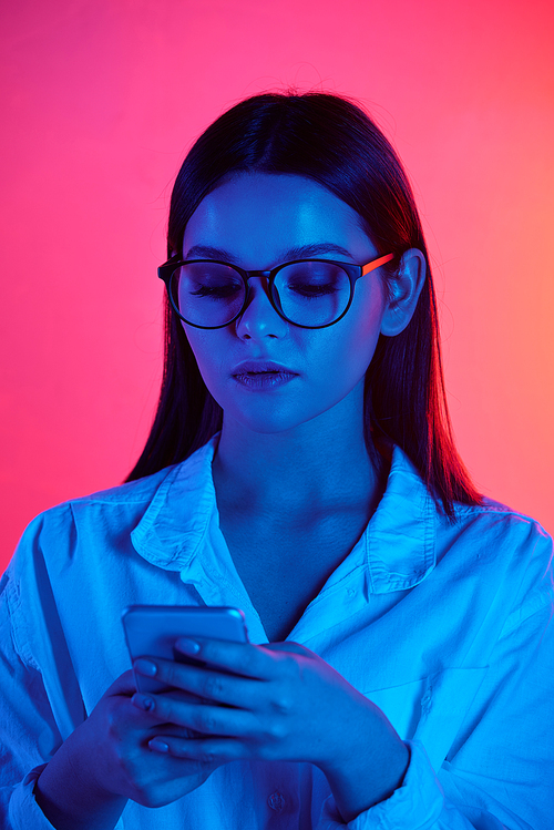 Serious female office worker in eyeglasses and white shirt using mobile phone in isolation on pink neon background