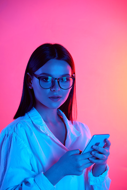 Portrait of modern brunette girl in glasses using smartphone in neon blue light against pink background