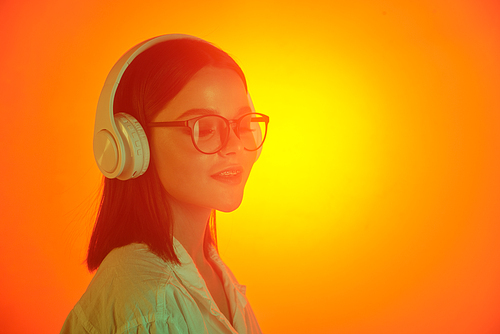 Young woman in eyeglasses and white shirt enjoying her favorite music in headphones over orange neon background