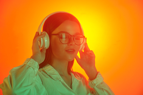 Happy relaxed young woman in wireless headphones and glasses listening to music with closed eyes against bright orange background