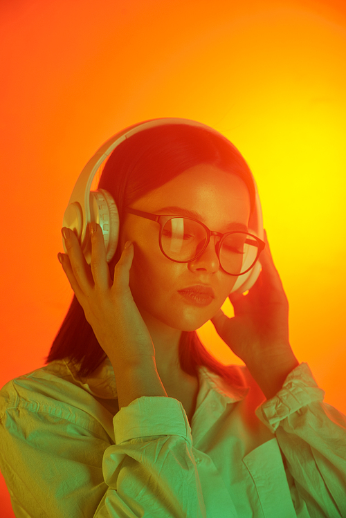 Pretty serene brunette female in white headphones enjoying relaxing music over orange neon background in isolation