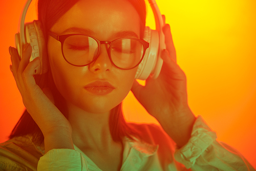Close-up of calm young woman in eyeglasses holding headphones while listening to music in neon light