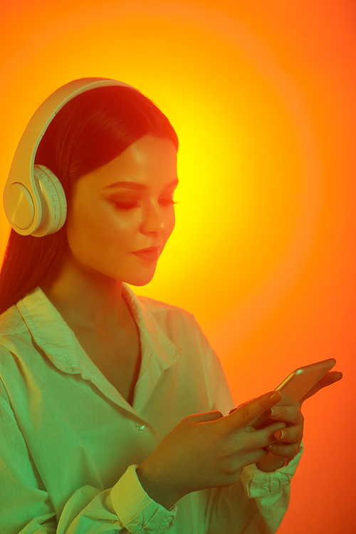 Content brunette girl in wireless headphones choosing music track on smartphone against bright orange background