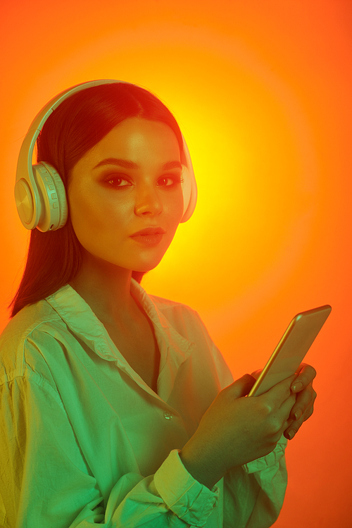 Portrait of content young woman in wireless headphones using modern phone against orange background