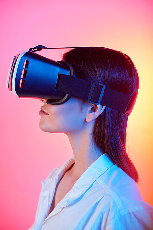 Side view of brunette girl in white blouse using virtual reality goggles against tender pink and yellow background