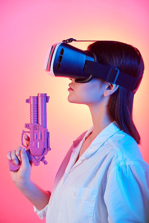 Pretty girl in vr headset holding violet plastic gun while shooting at virtual target against pink neon background in isolation