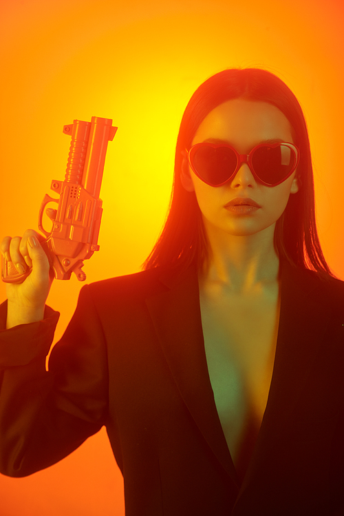Portrait of serious sexy girl in jacket and heart-shaped sunglasses raising gun against orange background