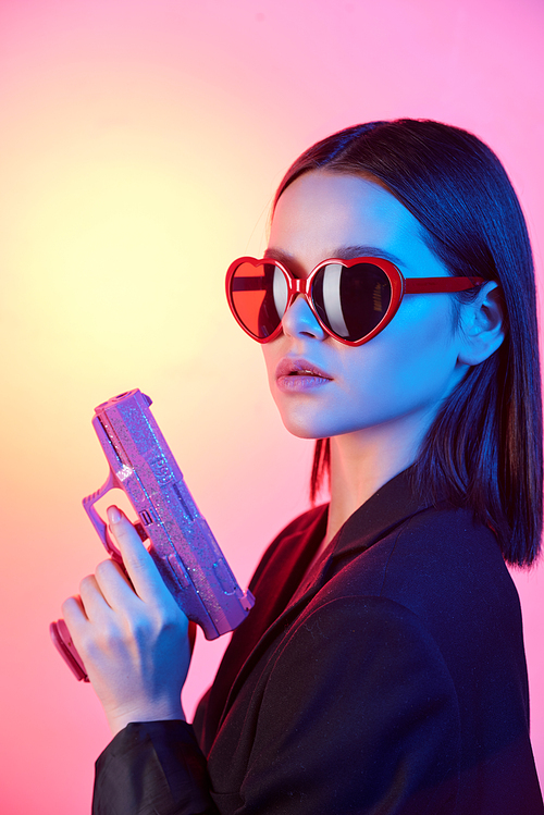 Young luxurious brunette woman in heart-shaped sunglasses and black jacket holding violet plastic gun against neon background