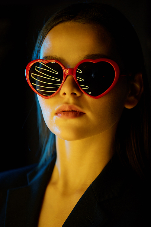 Young attractive beunette woman in heartshaped sunglasses and dark formalwear standing in front of camera in isolation