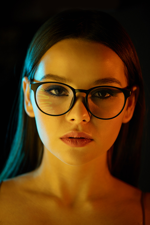 Portrait of serious young brunette woman in glasses against dark background, nightlife or clubbing concept