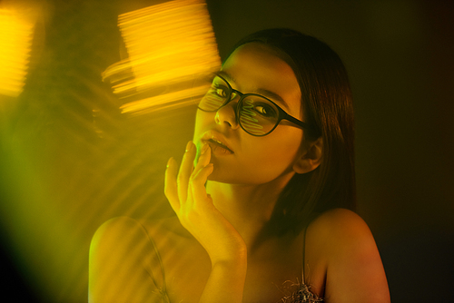 Young attractive female in eyeglasses sitting by window in front of camera and looking at you on neon background