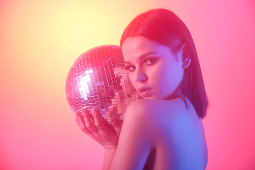 Portrait of serious naked girl holding disco mirror ball against light yellow and pink background