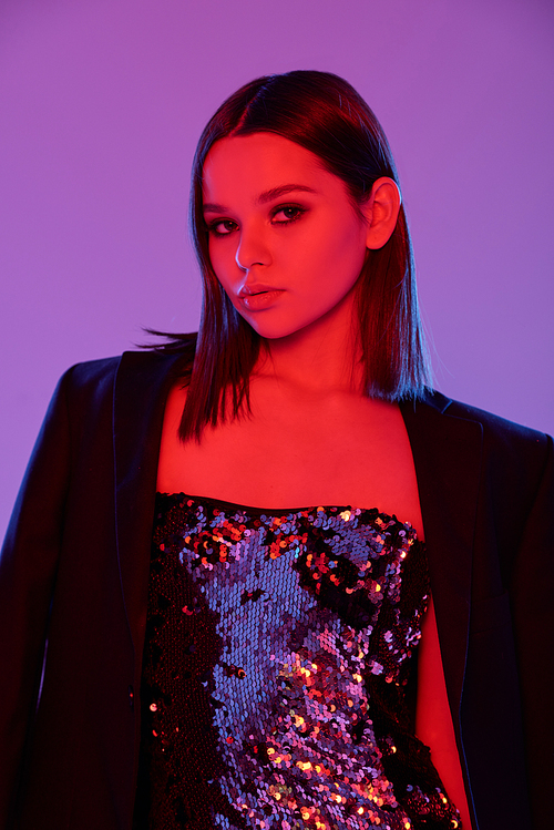 Pretty young brunette in little glittering dress and black jacket standing against violet neon background in isolation