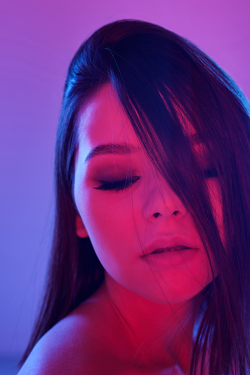 Young elegant female with dark long hair and perfect makeup posing in front of camera on violet neon background