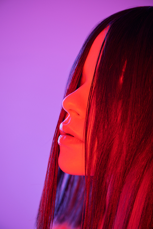 Face of young luxurious woman with dark long smooth hair standing against violet neon background in isolation