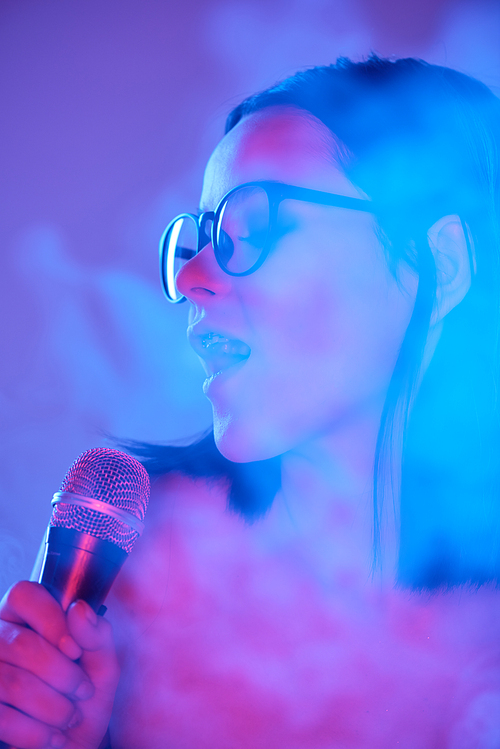 Pretty young emotional female singer with microphone by mouth performing songs in smoke against neon background