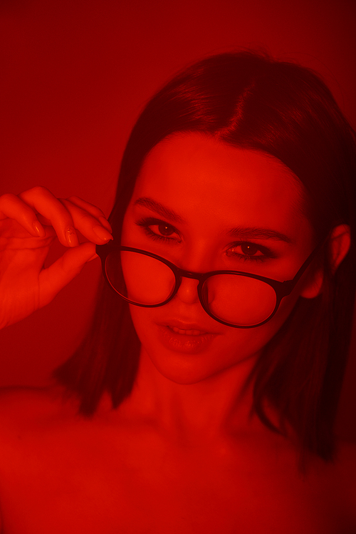 Young elegant pretty woman with eyeglasses looking at you in isolation through red neon light in the night club
