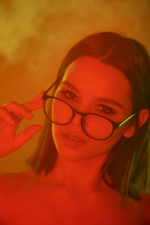 Portrait of ravishing young lady with brown hair adjusting glasses in smoky room