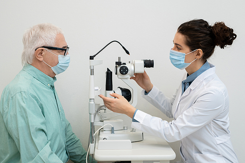 Side view of young female ophthalmologist using medical equipment in clinics while testing eyesight of senior male patient in protective mask