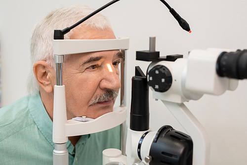 Senior male patient with white hair sitting in front of ophthalmological equipment while having his eyesight checked in contemporary clinics