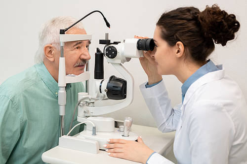 Young brunette female ophthalmologist in whitecoat using medical equipment in clinics while testing eyesight of senior male patient