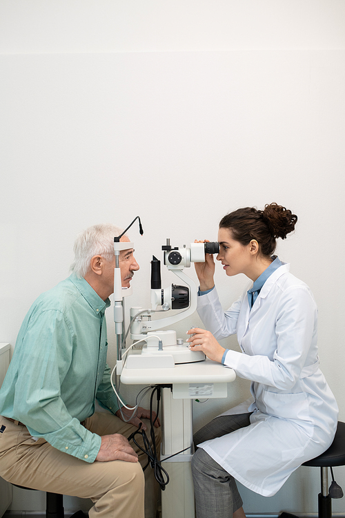 Side view of young brunette female ophthalmologist in whitecoat using medical equipment in clinics while checking up eyesight of senior man