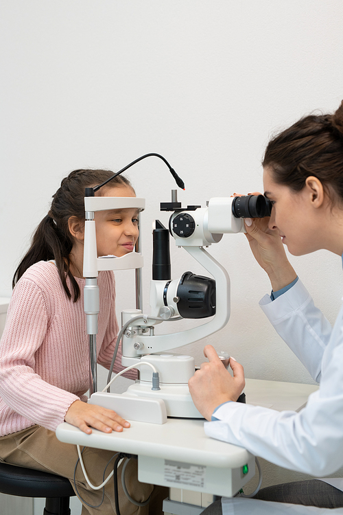 Young brunette female ophthalmologist in whitecoat using medical equipment while testing eyesight of cute little girl in modern clinics