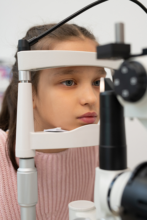 Adorable elementary schoolgirl sitting in front of ophthalmological equipment while having her eyesight checked in contemporary clinics
