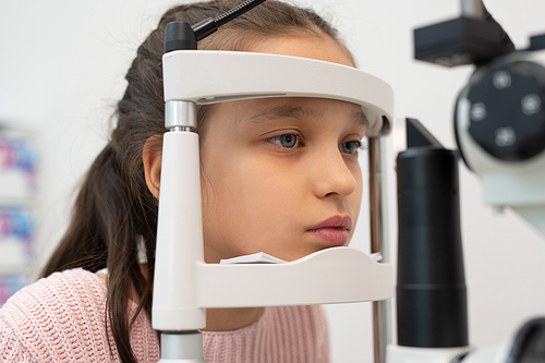 Face of elementary schoolgirl sitting in front of ophthalmological equipment while having her eyesight checked in modern clinics