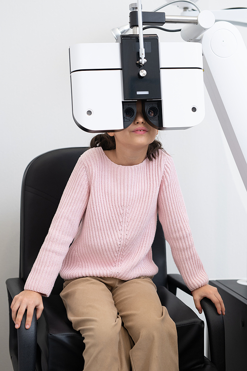 Elementary schoolgirl in casualwear sitting on chair in front of ophthalmological equipment while having her eyesight checked up