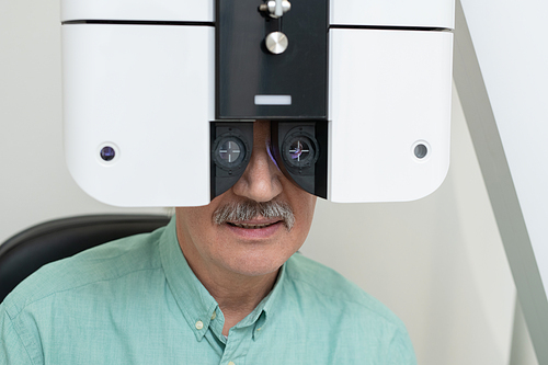 Face of senior male patient sitting in front of ophthalmological equipment while having his eyesight checked up in contemporary clinics