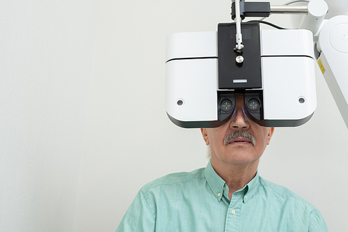 Senior male patient sitting in front of ophthalmological equipment and looking through lens while having his eyesight checked up in clinics