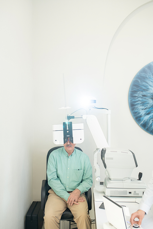 Senior male patient sitting on chair in front of ophthalmological equipment and looking through lens during medical check-up of his eyesight