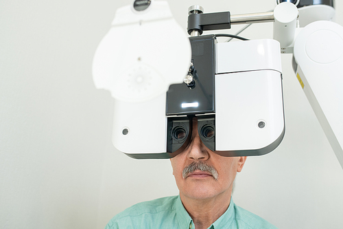 Senior male patient sitting in front of ophthalmological equipment and looking through lens during medical check-up of his eyesight in clinics