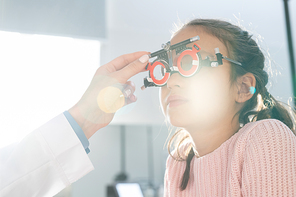 Elementary schoolgirl sitting in front of ophthalmologist during check-up of her eyesight with optometrist trial frame in contemporary clinics