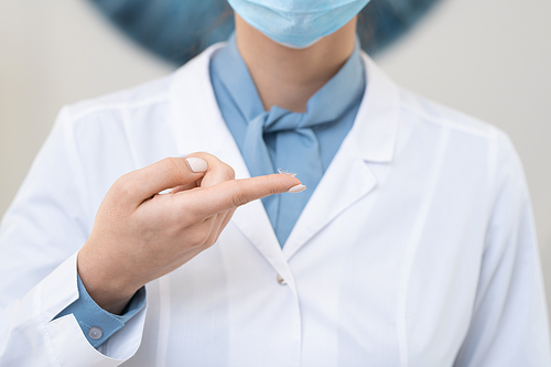 Hand of young contemporary female ophthalmologist in whitecoat and protective mask holding contact lens on forefinger in front of camera