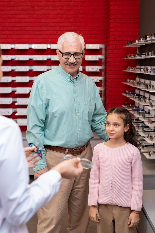 Happy senior man and his cute granddaughter standing in front of young female ophthalmologist in whitecoat offering them new eyeglasses