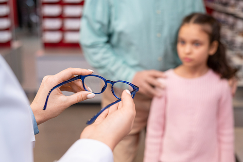 Hands of young female ophthalmologist in whitecoat offering pair of new eyeglasses to cute little girl visiting medical office with her grandfather