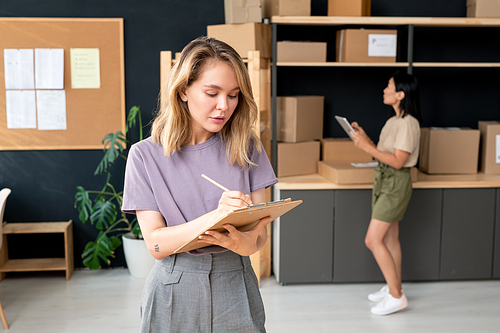 Serious young blond female manager of online trading office making notes in document while her colleague standing by shelves with packed orders