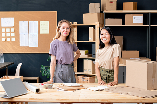 Young pretty intercultural workers of online shop office standing by wooden table in front of camera while working with orders