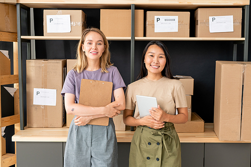 Two young cheerful workers of storage room of online shop with documents standing by large wooden shelves with packed boxes