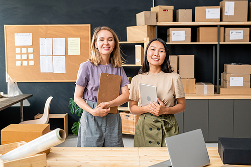 Two young cheerful female workers of online shop office standing by table against large wooden shelves with packed boxes and noticeboard