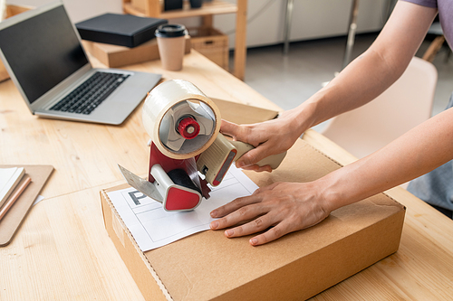Hands of female manager of online shop with cellotape gun dispenser sealing cardboard box with packed order of client by her workplace
