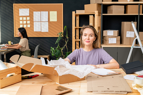 Young pretty female worker of online shop office sitting by table in front of camera, packing orders of clients and checking data in document