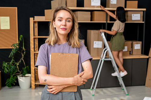 Young confident blond female worker of storage room with clipboard standing in front of camera against her colleague on stepladder