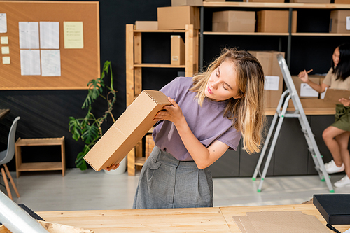 Young female manager with long blond hair checking quality of packing cardboard box with order of client while working in store-room
